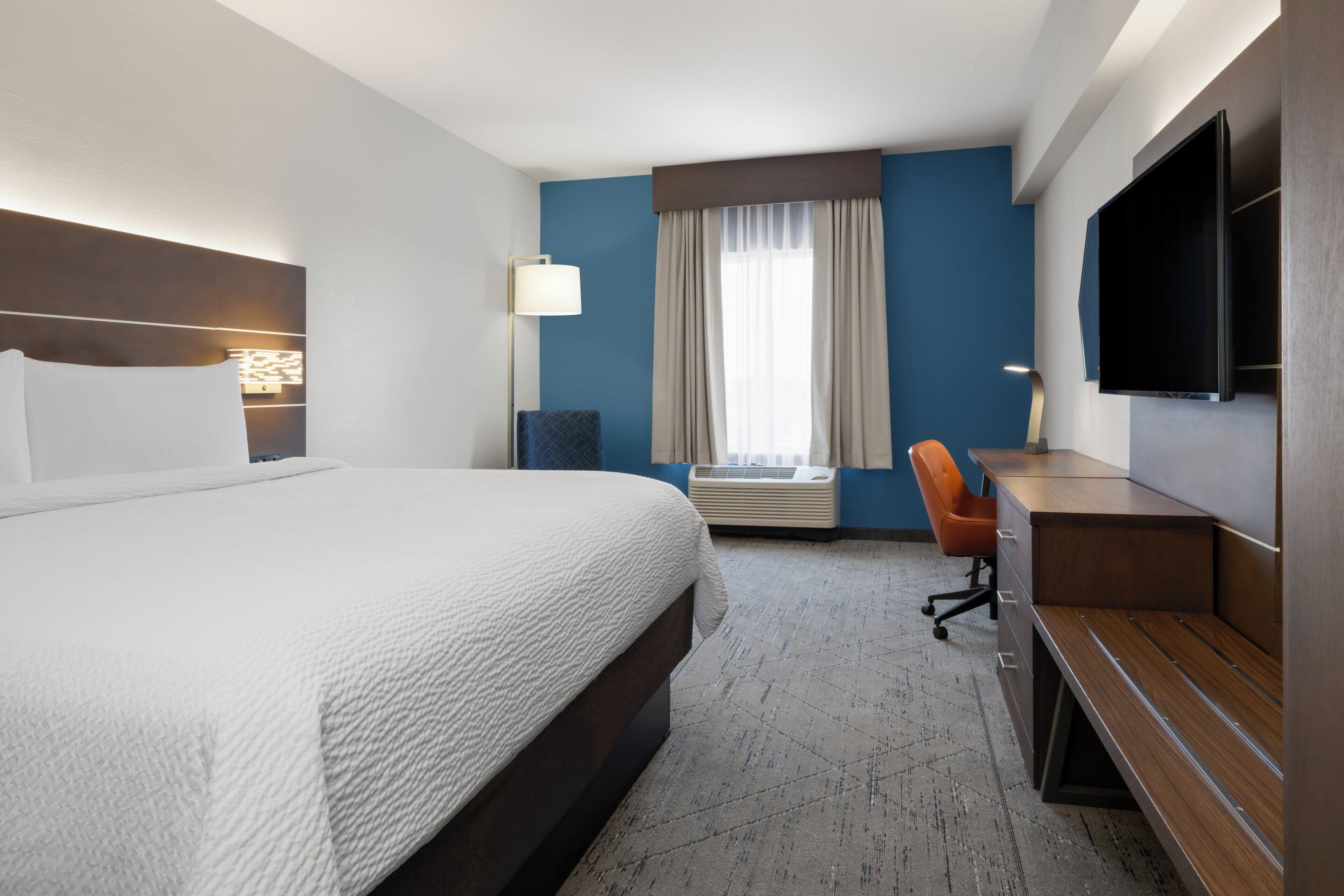 Habitación de hotel con una cama grande, un escritorio, una silla, un televisor de pantalla plana y un ventanal con cortinas.