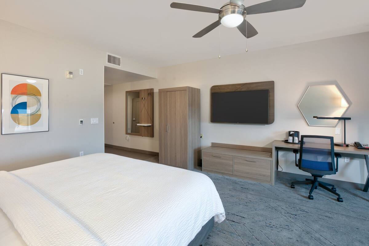 A hotel room with a bed, desk, chair, ceiling fan, artwork, and a TV.