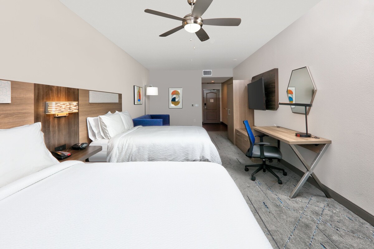 A hotel room with two beds, a desk, a chair, a TV, and a ceiling fan.