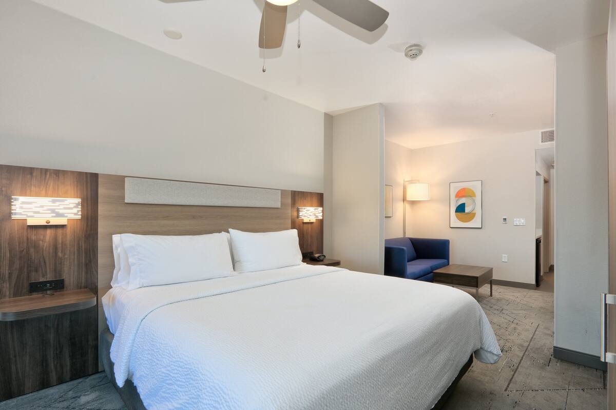 A modern hotel room with a large bed, wooden headboard, bedside lamps, a desk, and a blue chair.