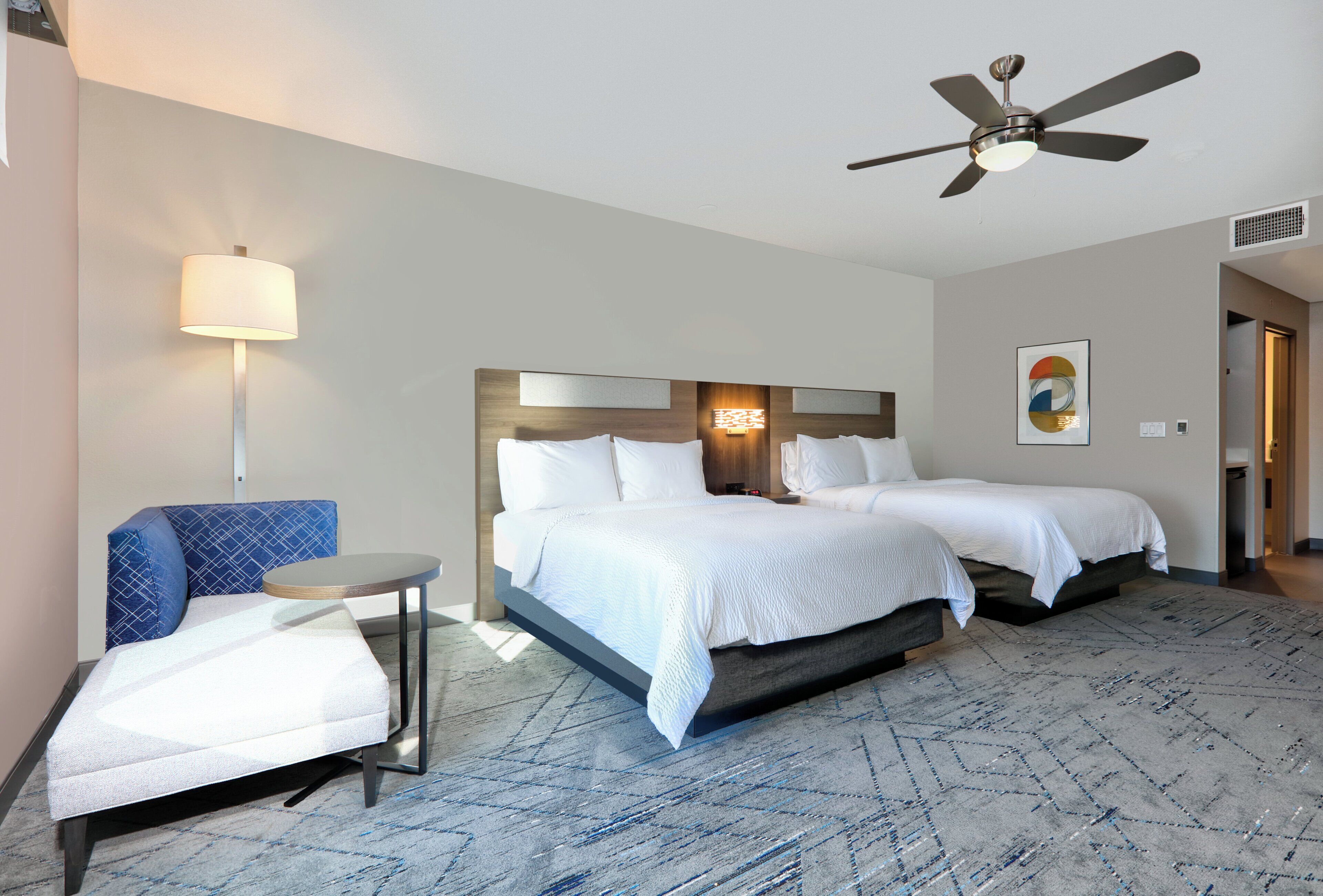 A hotel room with two beds, a desk, a chair, a ceiling fan, and a wall-mounted lamp.