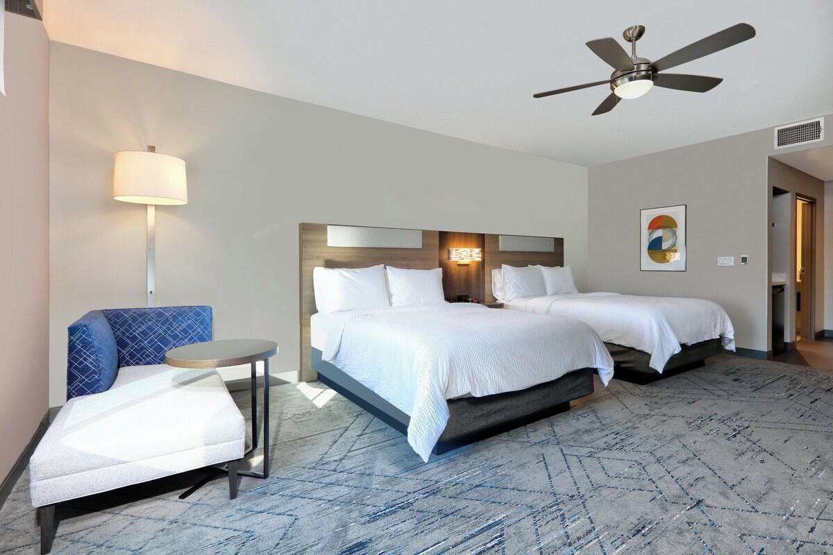 A hotel room with a bed, a chair, a small table, a ceiling fan, and a wall-mounted light.