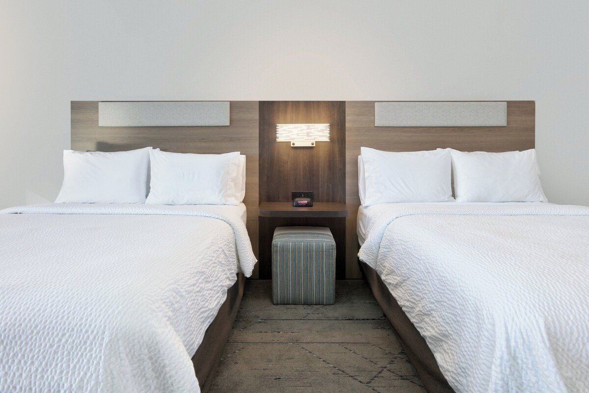 A hotel room with two single beds, a nightstand with a lamp, and a striped ottoman.
