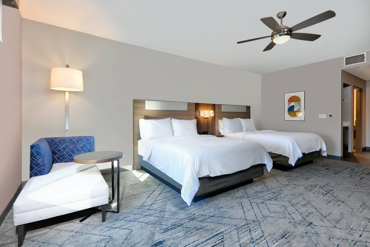 A hotel room with a bed, a chair, a small table, a ceiling fan, and a wall-mounted light.