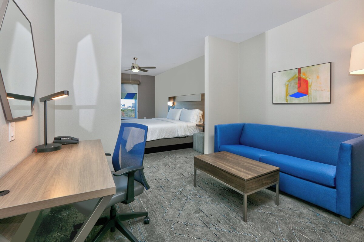 A hotel room with a bed, a blue sofa, a desk with a chair, a coffee table, a desk lamp, and a ceiling fan.