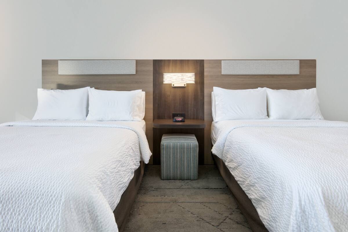 A hotel room with two single beds, a nightstand with a lamp, and a striped ottoman.