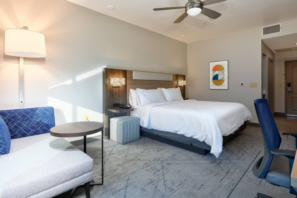 A modern hotel room with a bed, a sofa, a small table, and a ceiling fan.