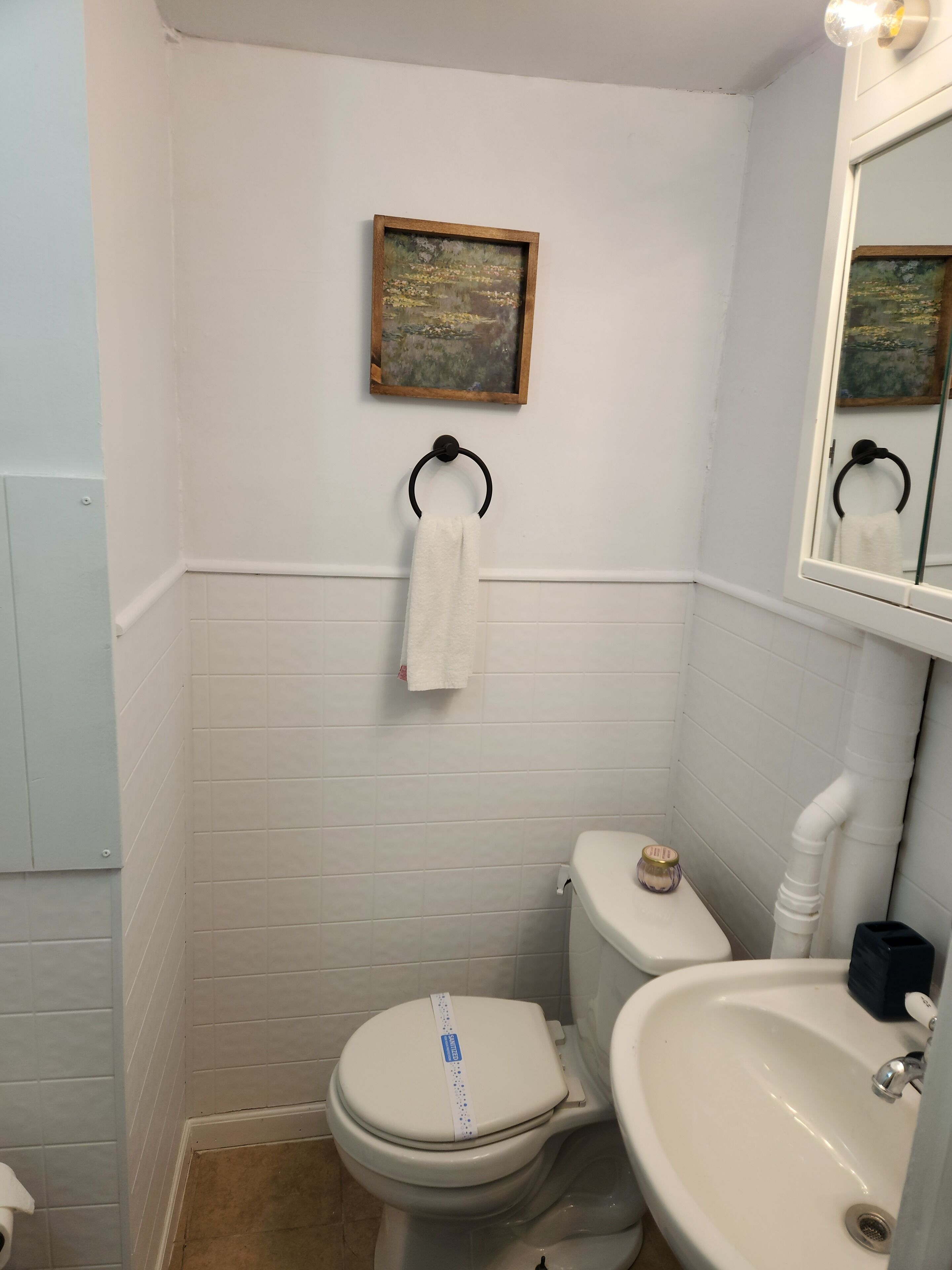A small, white-tiled bathroom with a toilet, a sink, and a framed painting on the wall.