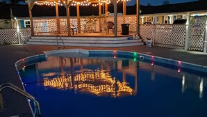 A swimming pool with a wooden deck and a covered seating area with string lights. - Jonathan Edwards Motel (Dennis Port)