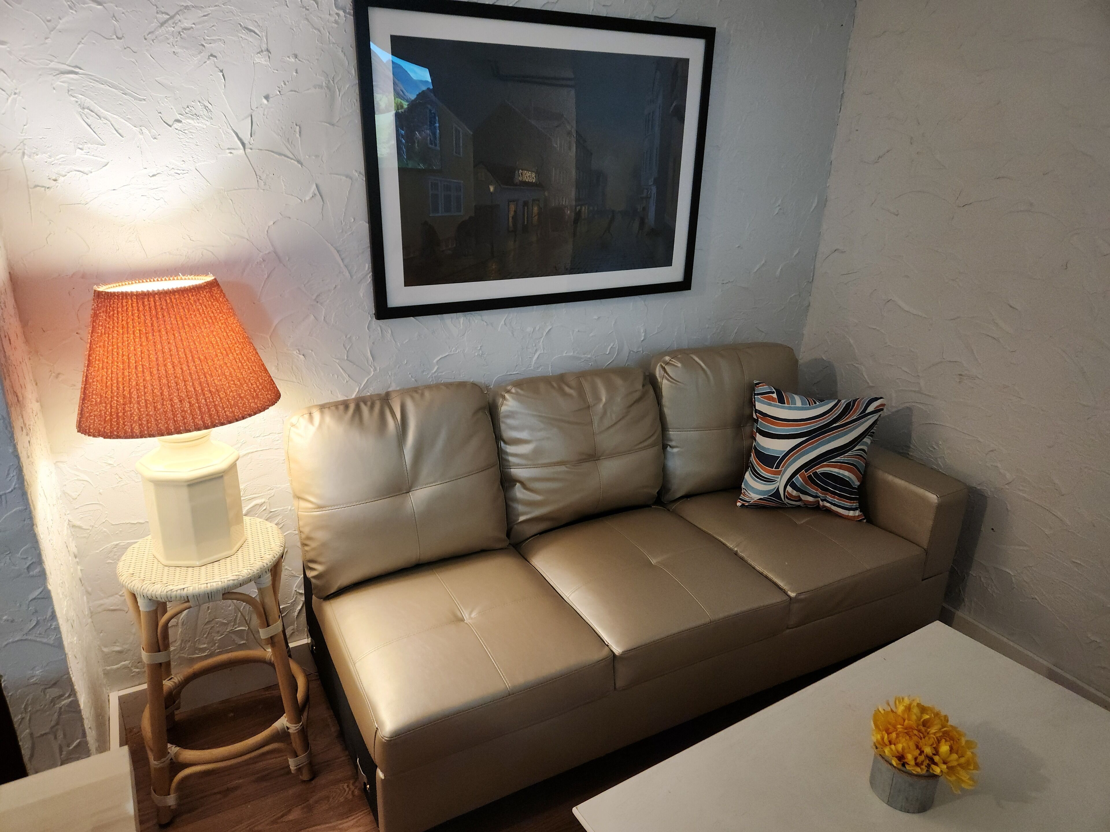A beige leather sofa with a patterned pillow, a wicker side table with a lamp, and a small vase with yellow flowers on a white table.