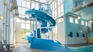 A large indoor swimming pool with a blue water slide, a sunlit area with tables and chairs, and a view of the outdoors through large windows. - Sandman Hotel & Suites Squamish (Squamish)