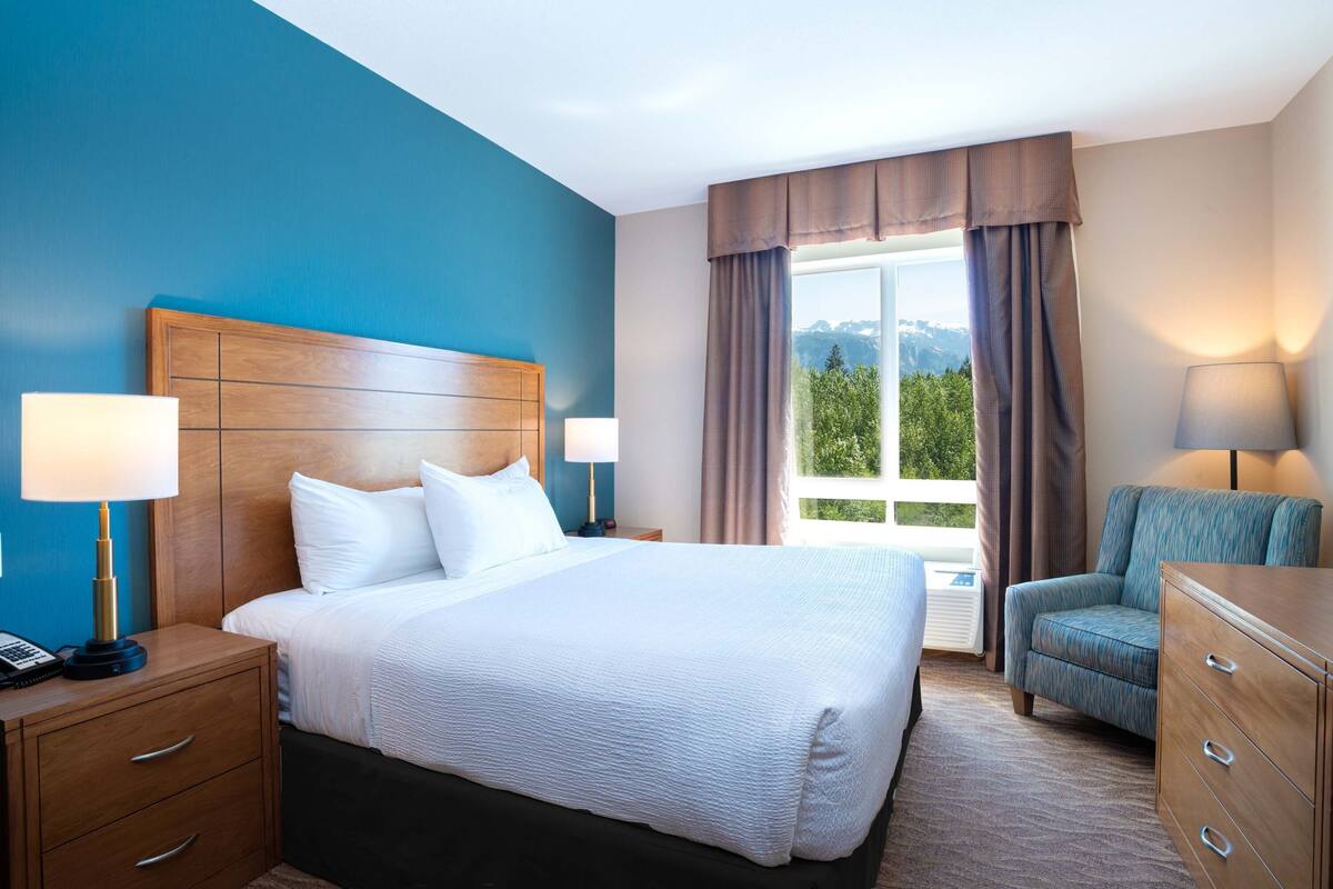 Una habitación de hotel con una cama grande, cabecero de madera, dos mesitas de noche con lámparas, una silla y un aparador.