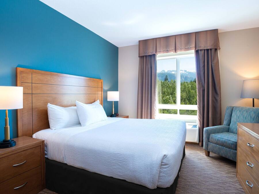 A hotel room with a large bed, wooden headboard, two bedside tables with lamps, a chair, and a dresser.