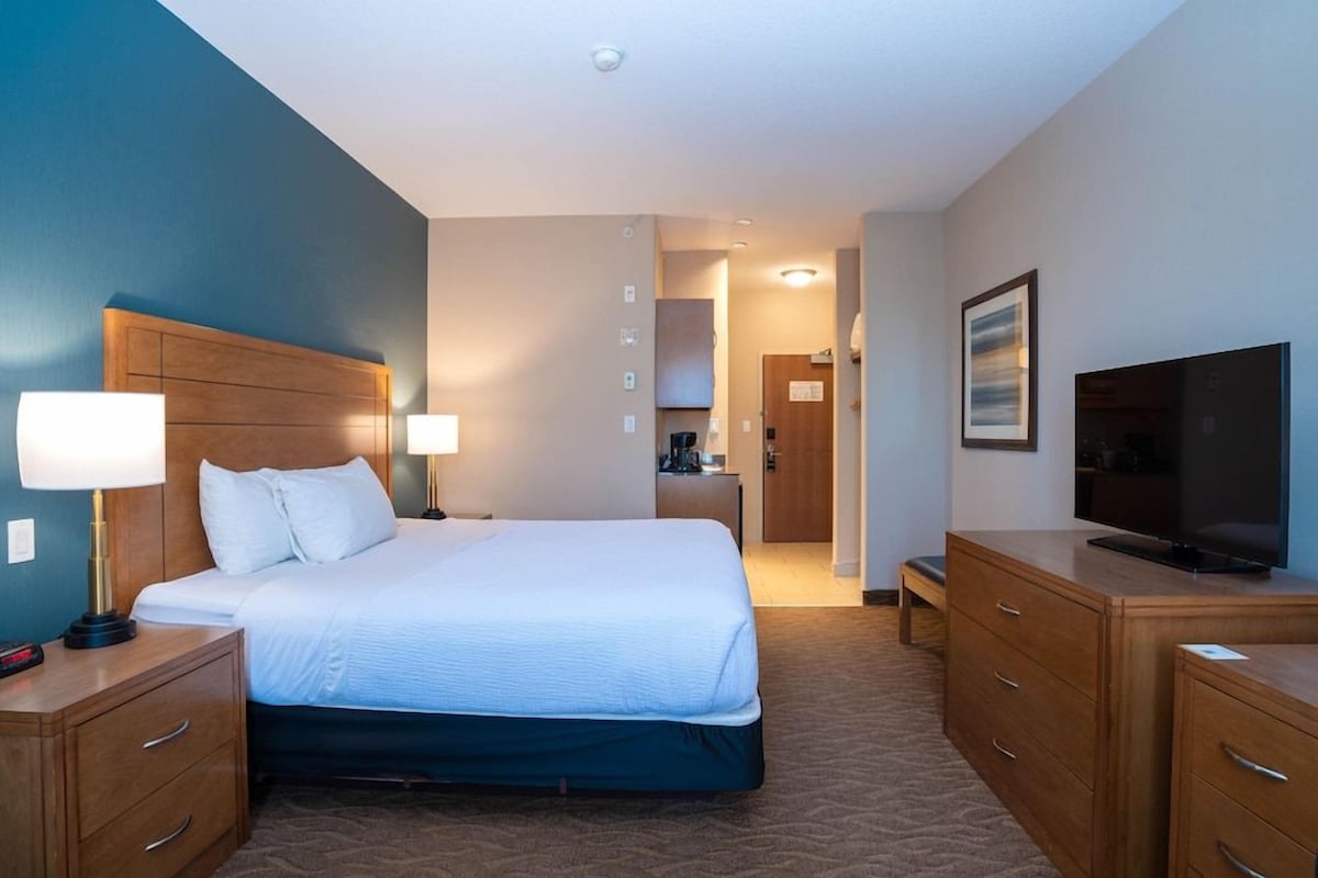 A hotel room with a large bed, wooden headboard, bedside tables with lamps, a flat-screen TV, and a view of the hallway.