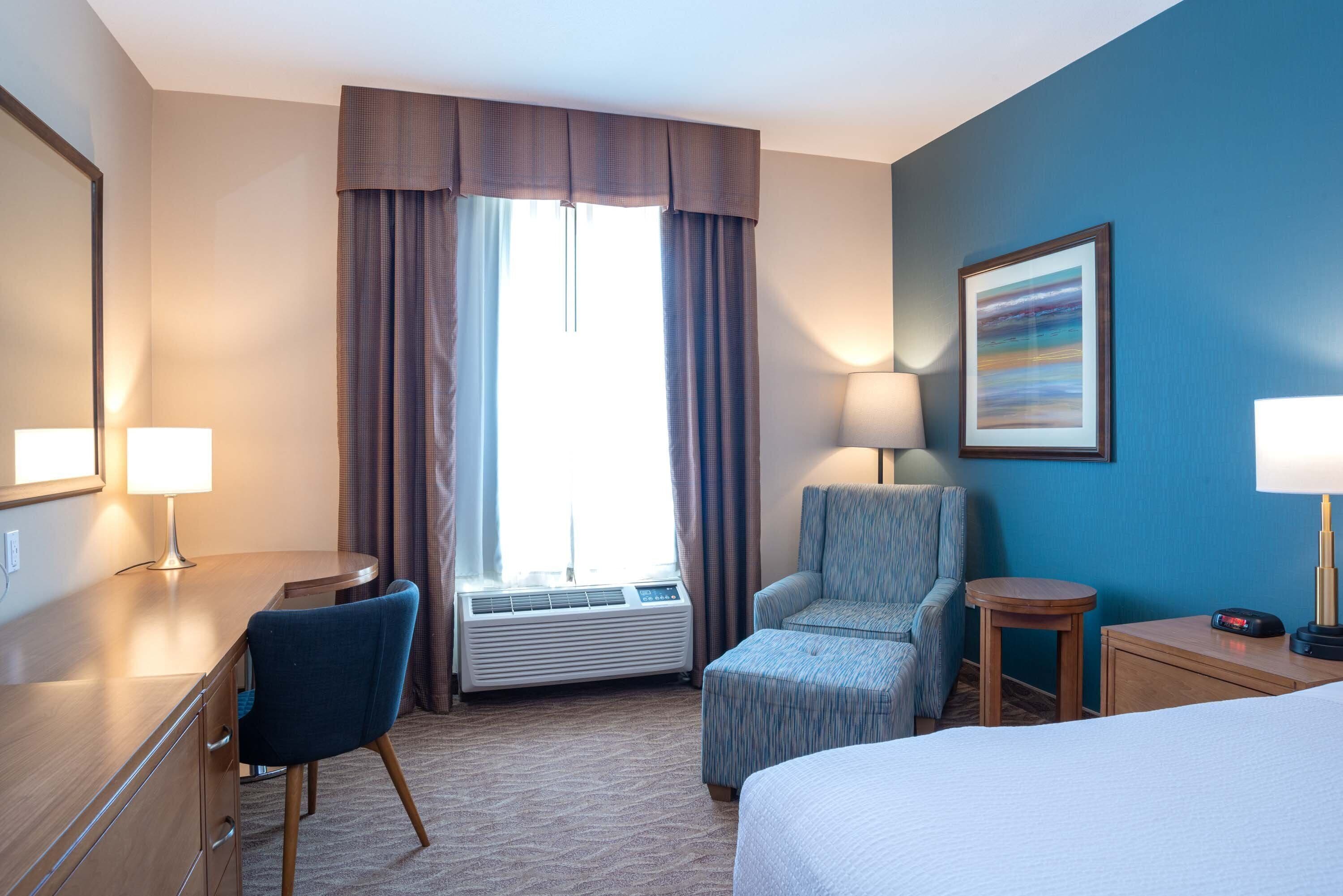 A hotel room with a bed, a desk with a chair, a small table, a standing lamp, a wall-mounted TV, and a window with curtains.
