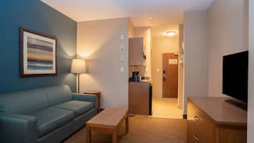 A modern hotel room with a sofa, a small table, a TV, a kitchen area, and a framed picture on the wall.