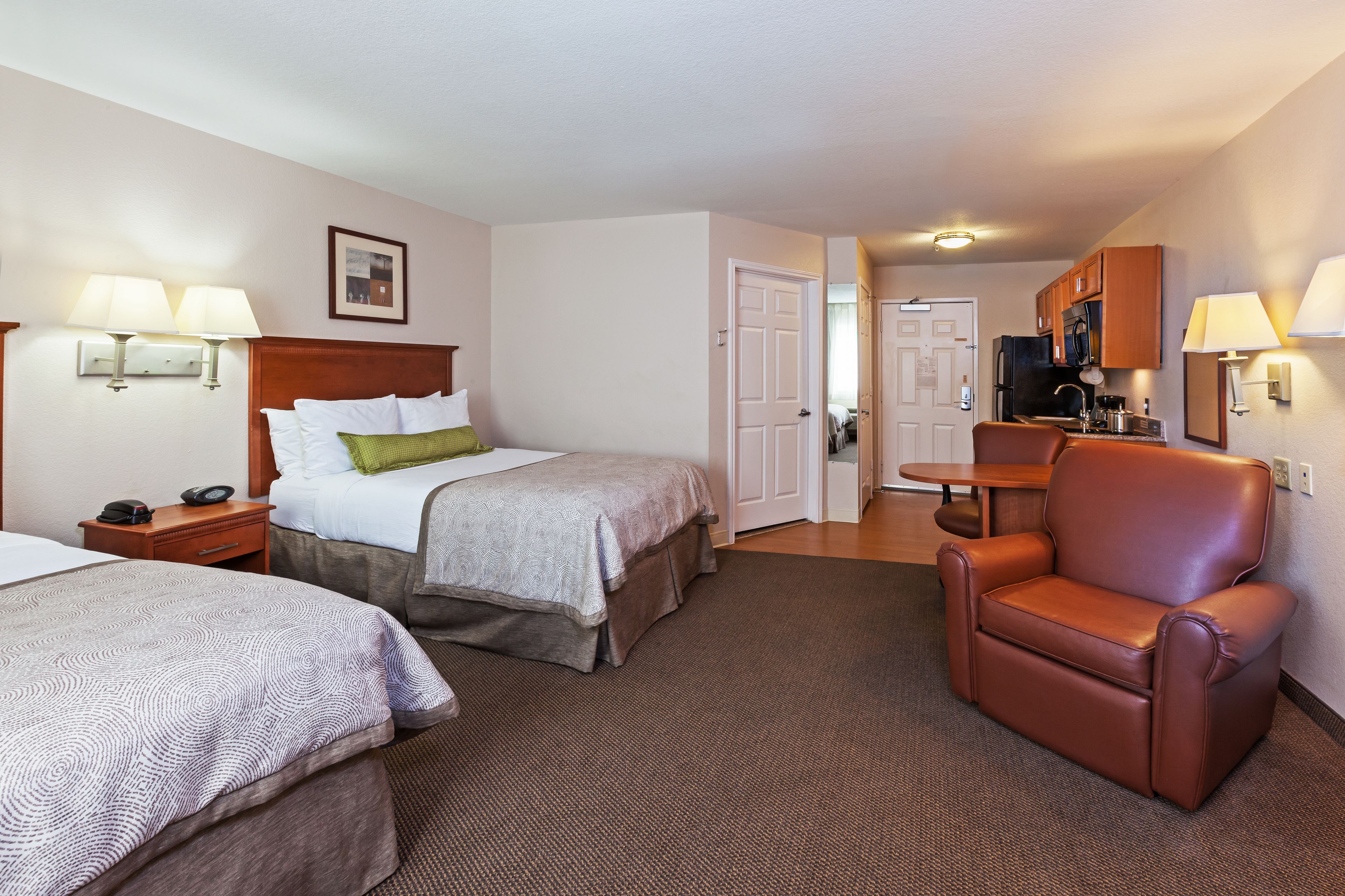 A hotel room with two beds, a brown leather chair, a kitchenette, and a door leading to another room.