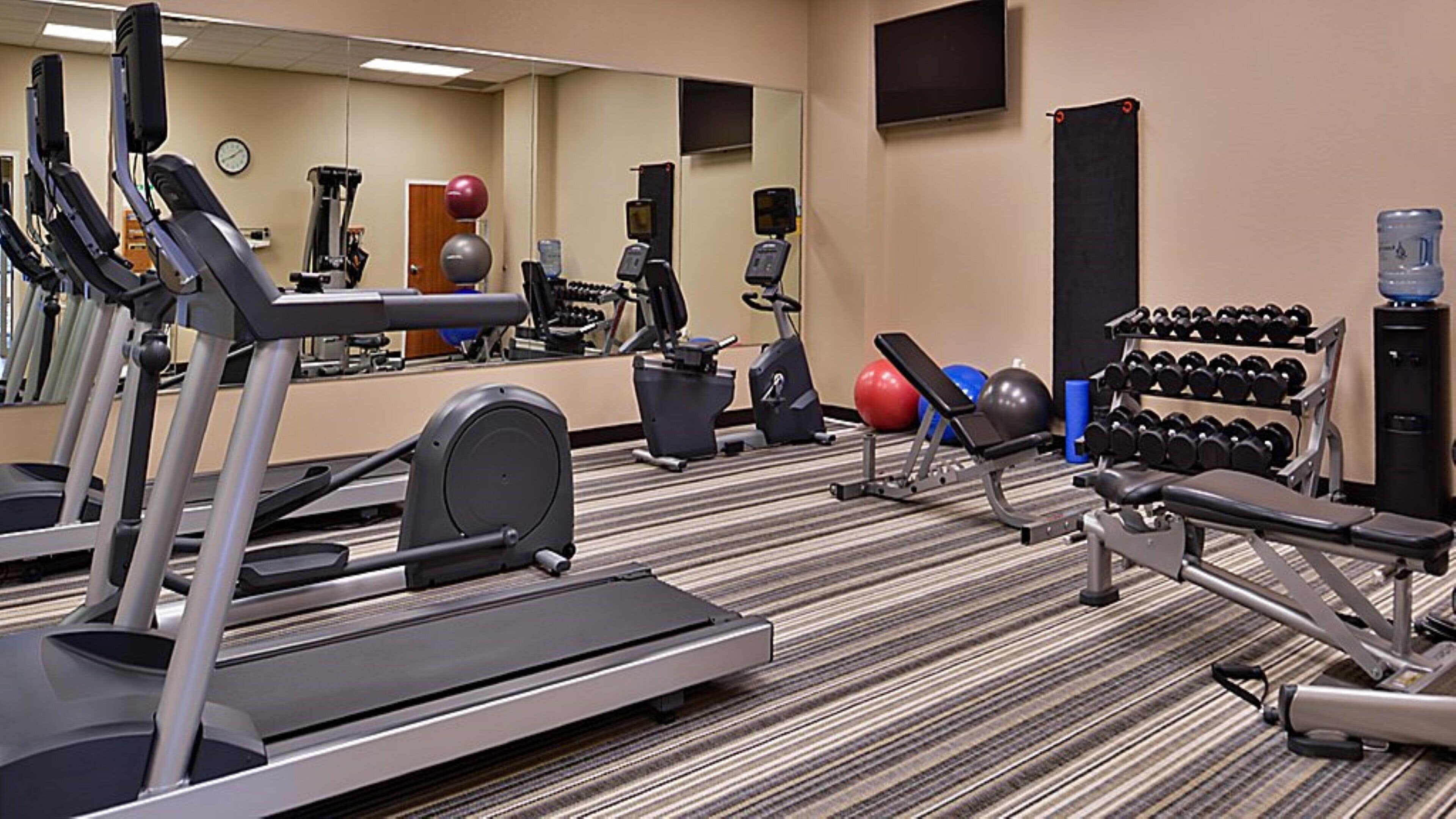 A hotel gym with treadmills, elliptical machines, a bench press, and a rack of dumbbells.