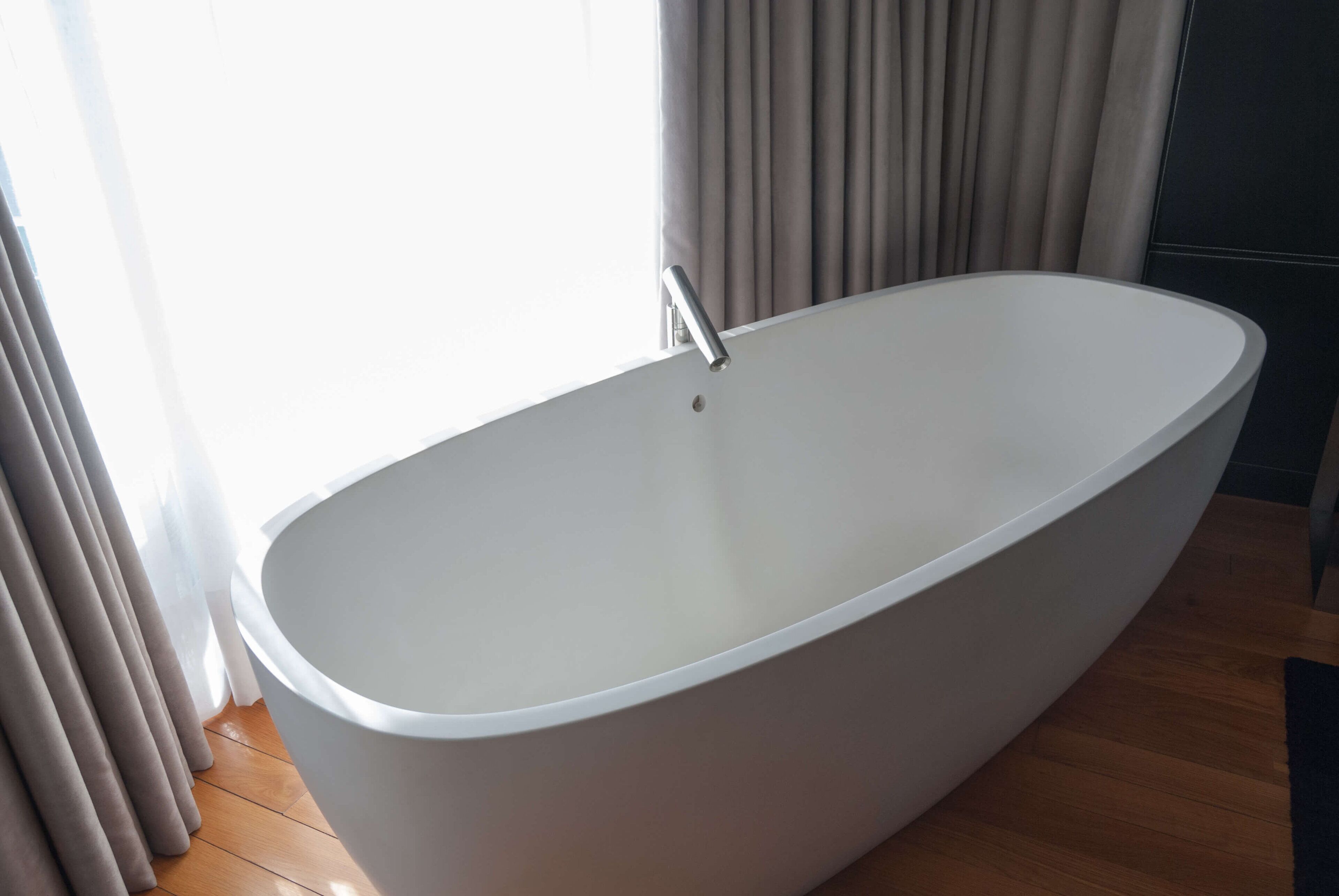 Een moderne badkamer met een vrijstaand bad, houten vloer en een raam met gordijnen.