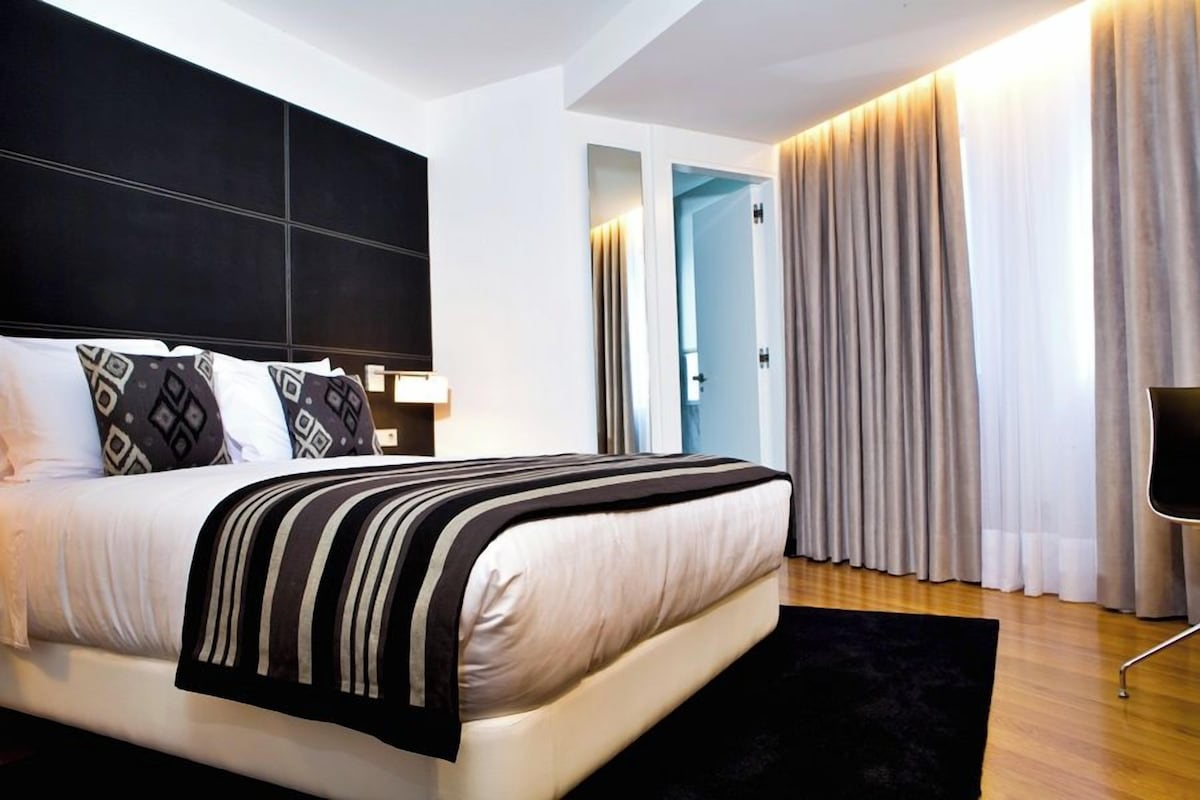 A modern hotel room with a large bed, a black and white striped throw, a black floor lamp, and a view of the outside through a door.
