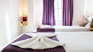 A hotel room with two beds, purple bedding, a white blanket, a TV on the wall, and a window with purple curtains. - Lancaster Court Hotel (London)