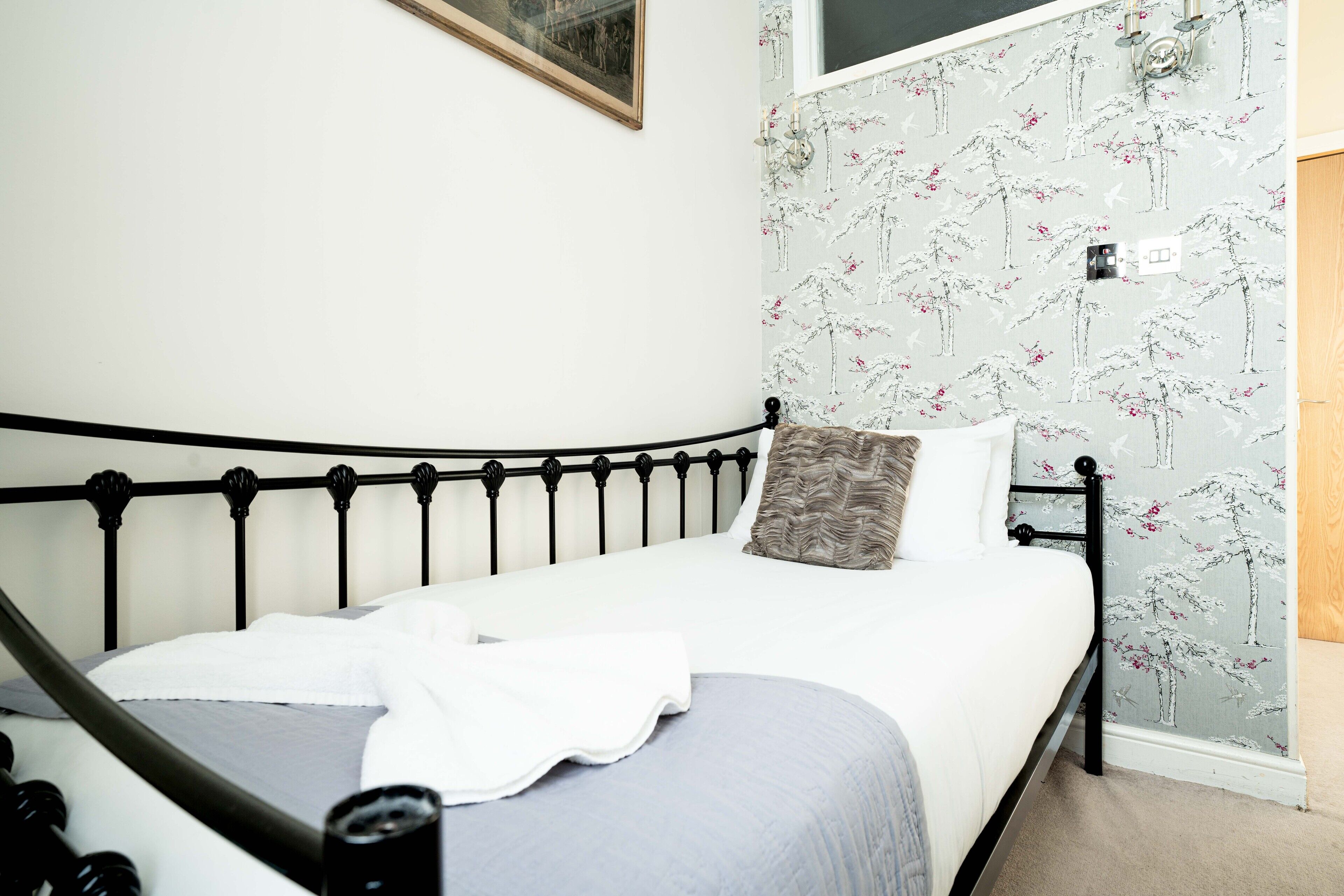 A neatly made bed with a metal frame, white bedding, and a floral wallpaper pattern on the wall behind it.