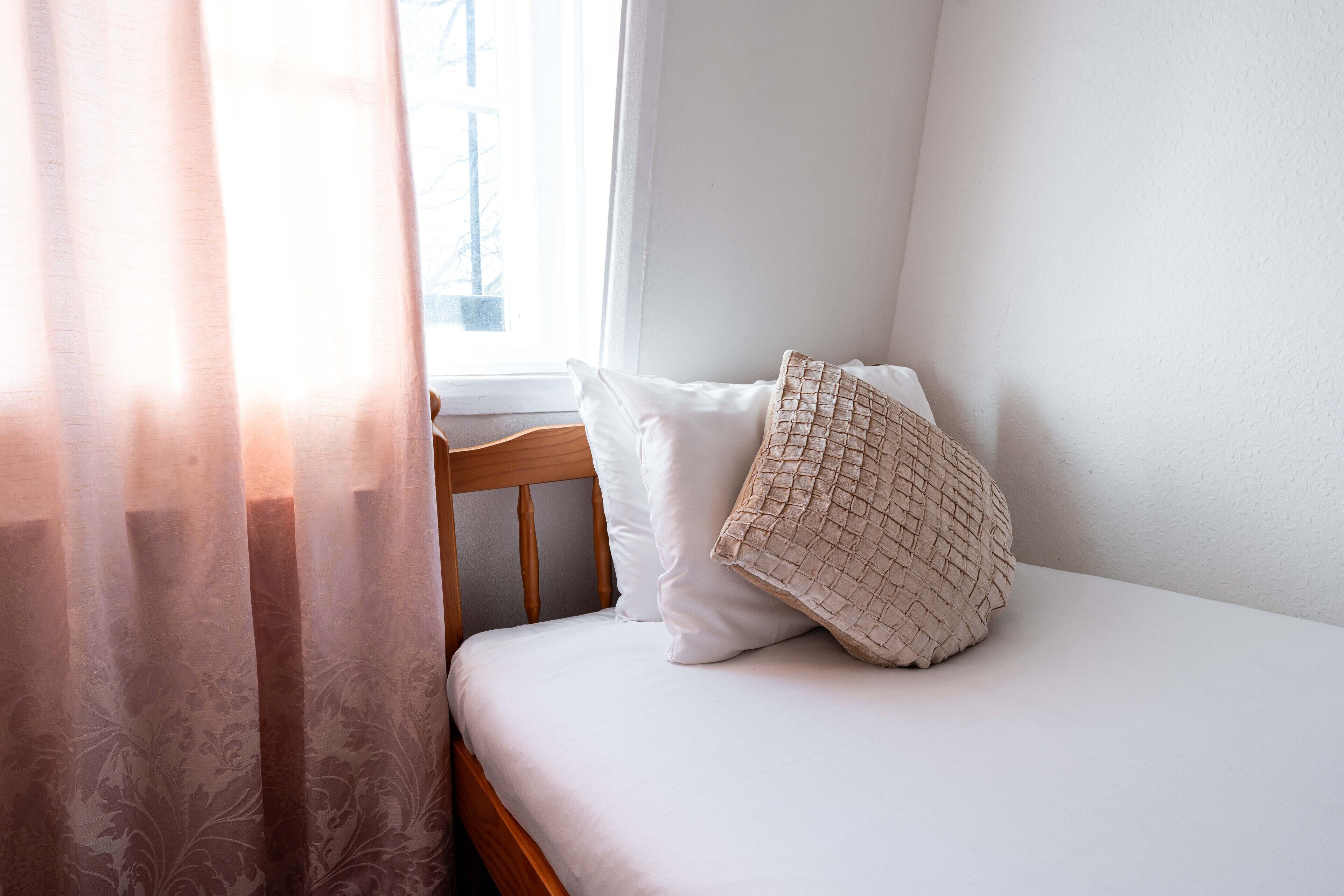 A neatly made bed with white linens and a woven throw pillow, a wooden headboard, and a window with sheer curtains.