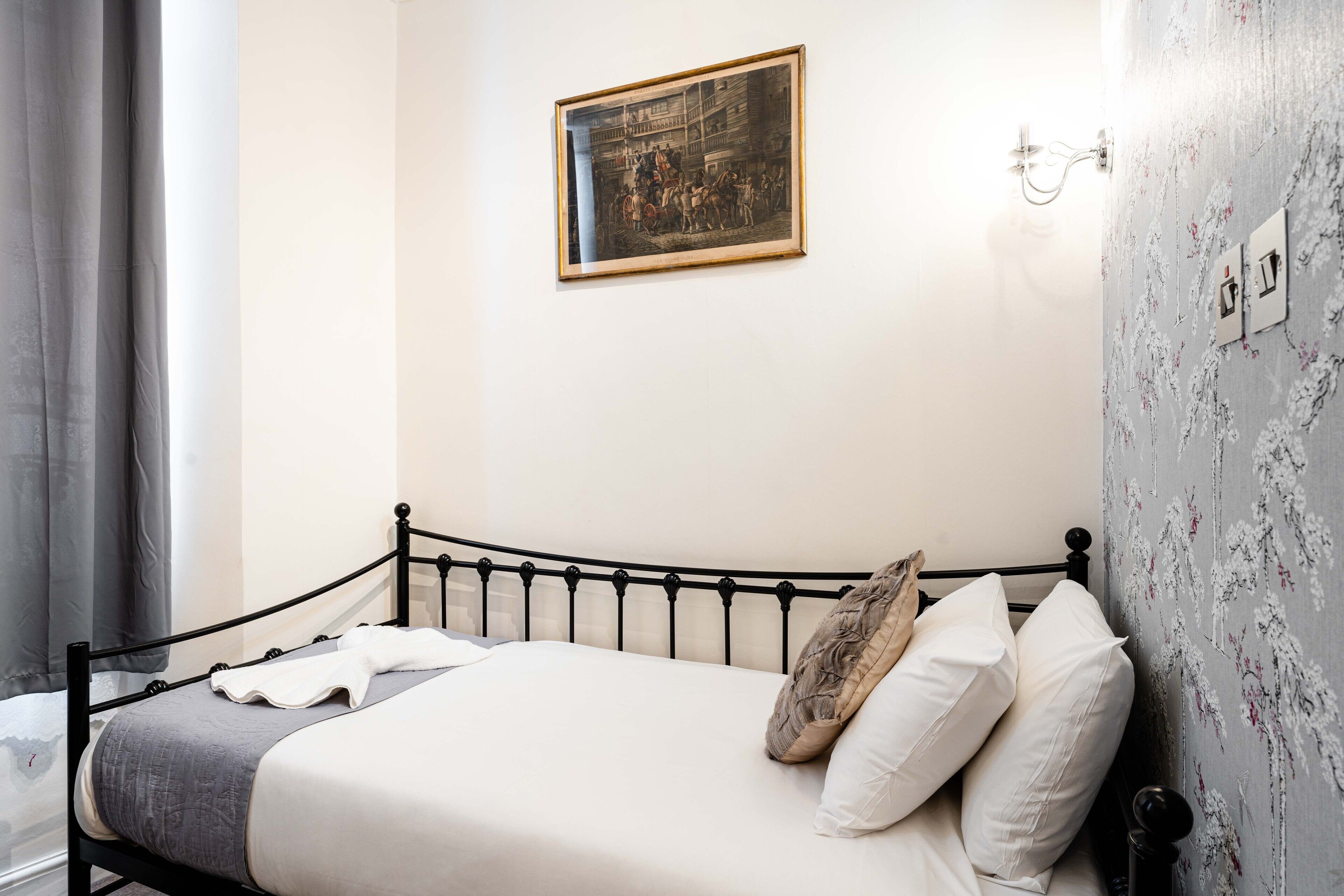 A neatly made bed with a headboard, a framed picture on the wall, and a floral wallpaper pattern.