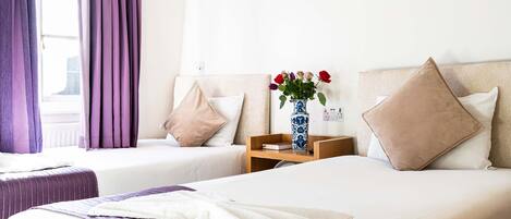 A hotel room with two beds, a nightstand with a vase of flowers, and a window with purple curtains.