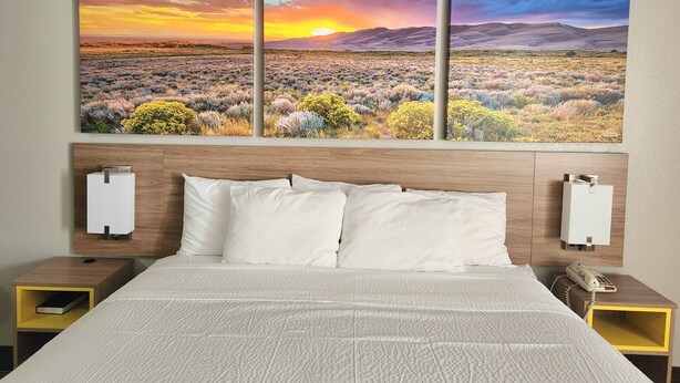 A hotel room with a large bed, white bedding, a wooden headboard, and a wall-mounted painting of a desert landscape. - Days Inn by Wyndham Lumberton (Lumberton)