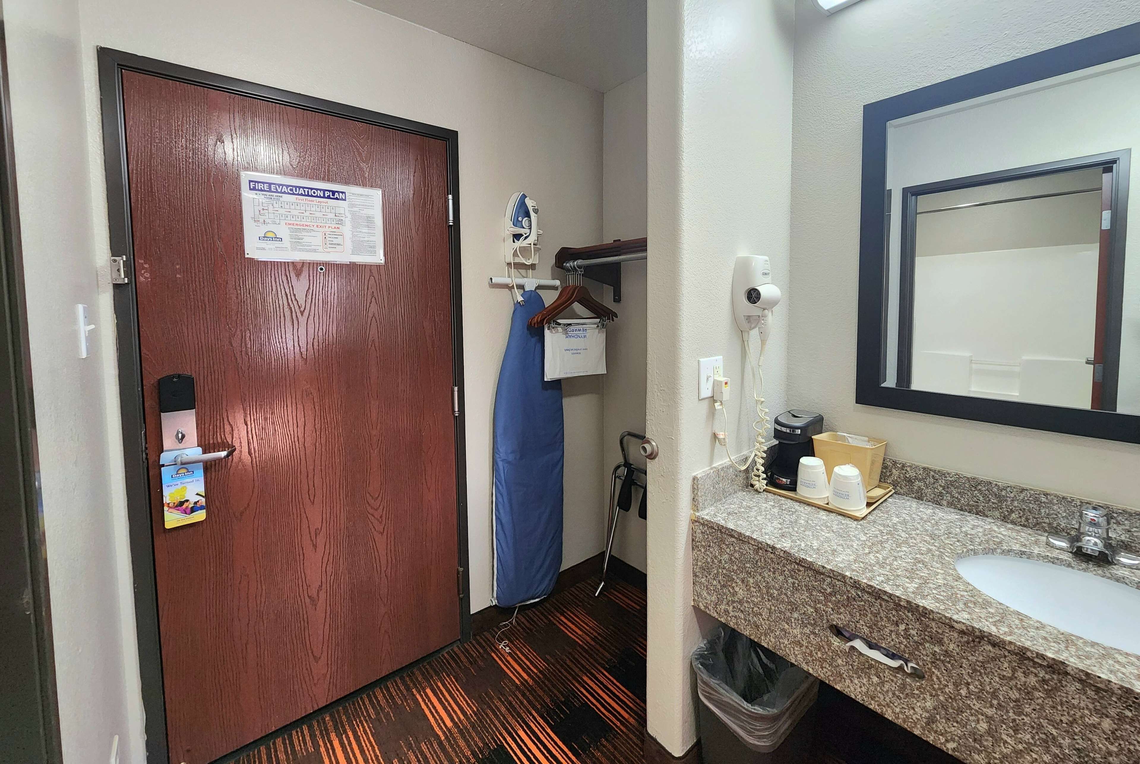 A hotel room with a wooden door, a fire evacuation plan, a blue curtain, a hairdryer, a mirror, a sink, and toiletries.