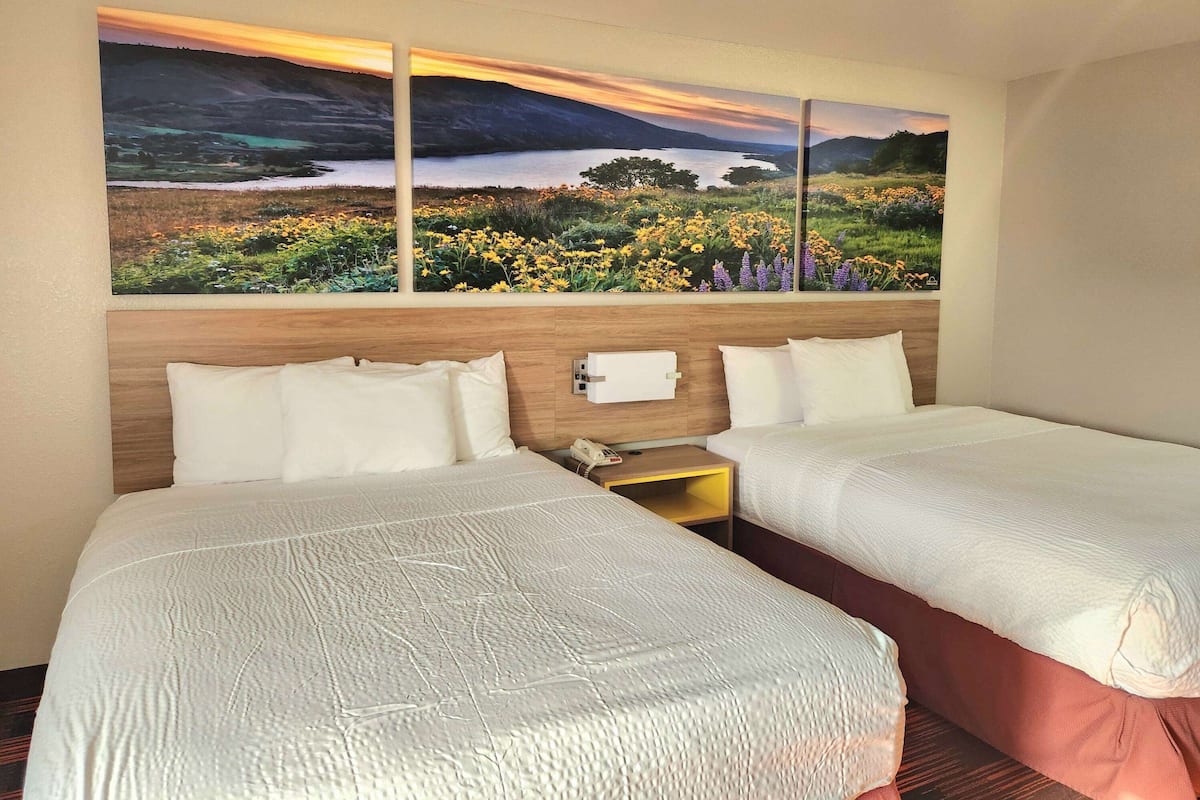 A hotel room with two beds, a wooden headboard, and a large landscape painting above the beds.