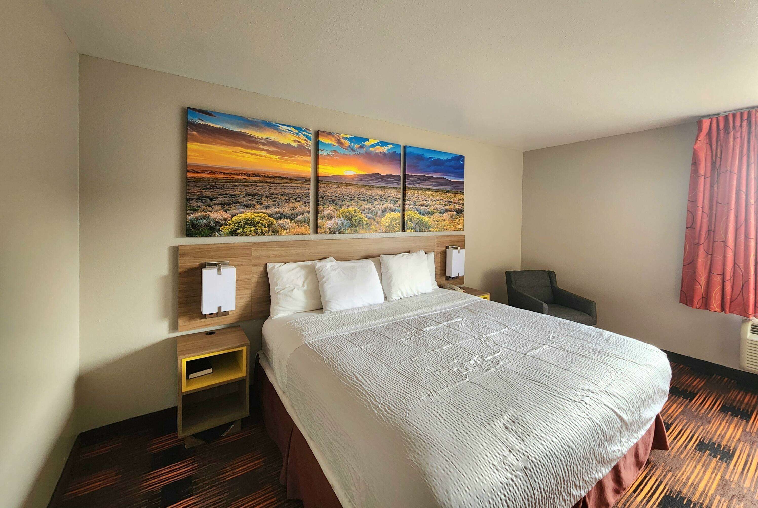 A hotel room with a large bed, a nightstand, a desk, and a painting of a sunset landscape on the wall.