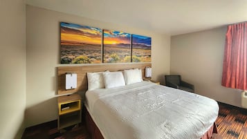 A hotel room with a large bed, a nightstand, a desk, and a painting of a sunset landscape on the wall.