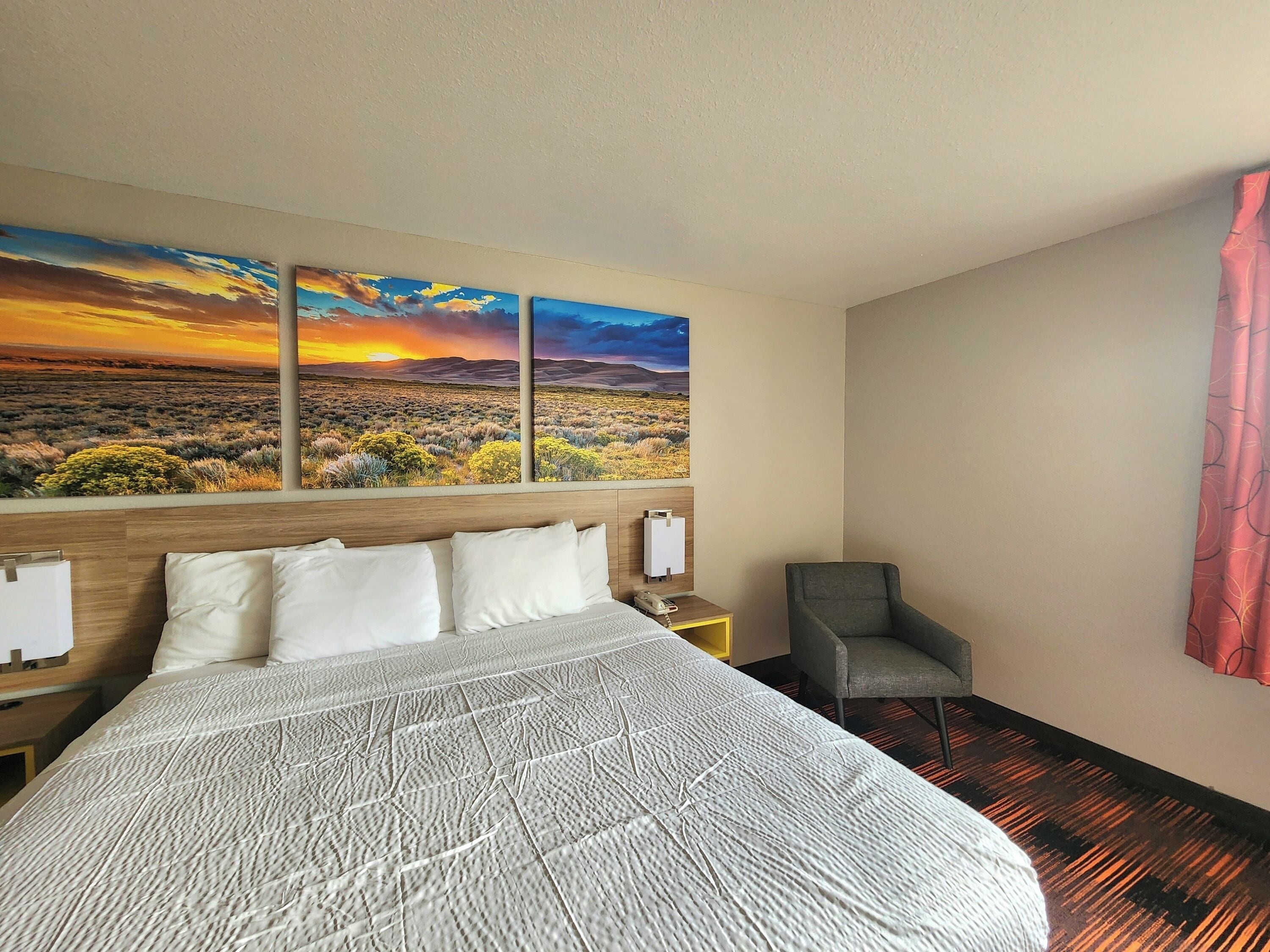 A hotel room with a large bed, a chair, a nightstand, and a wall with a sunset landscape painting.