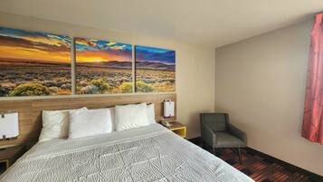A hotel room with a large bed, a chair, a nightstand, and a wall with a sunset landscape painting.