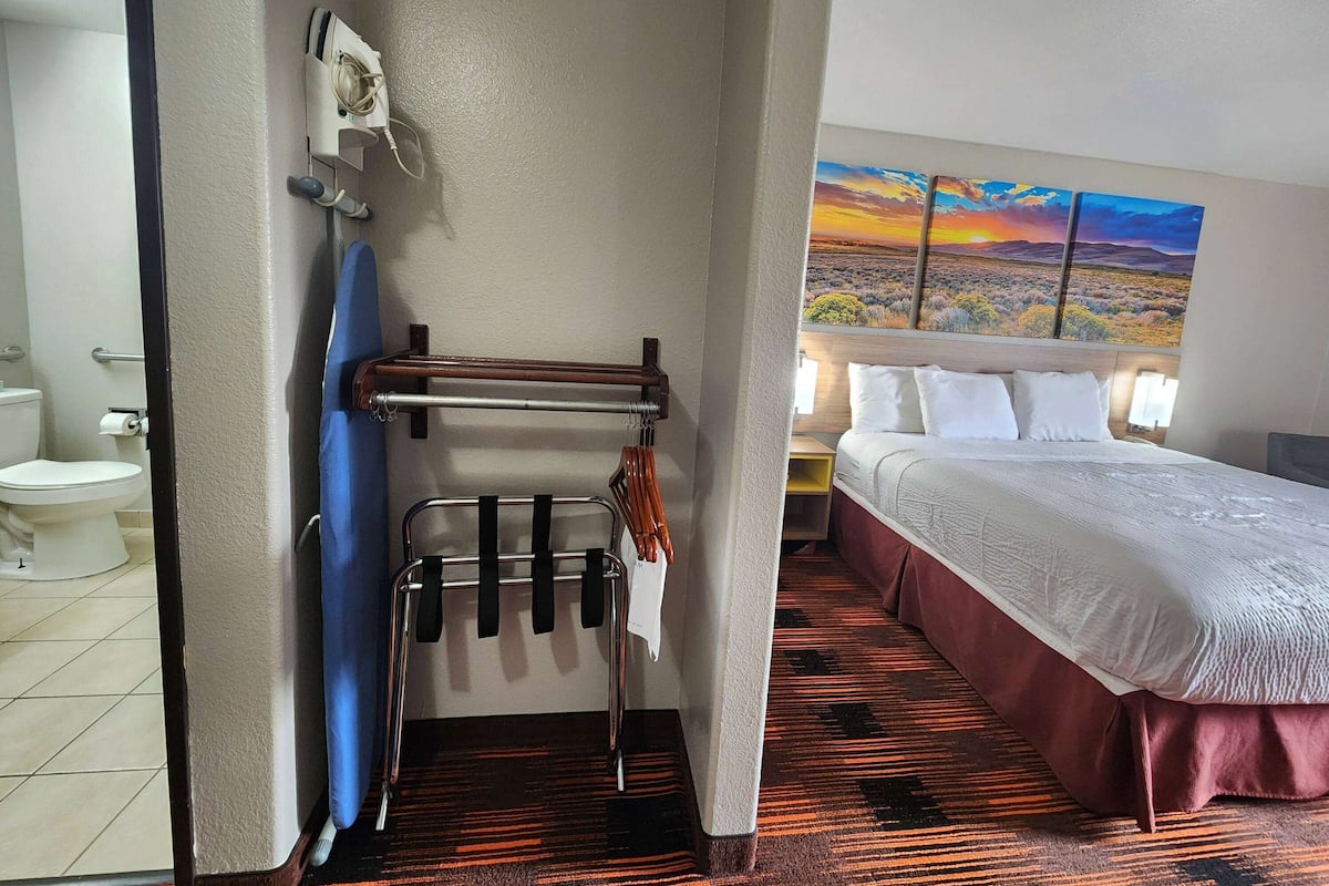 A hotel room with a bed, a painting of a sunset, a bathroom with a toilet and towel rack.