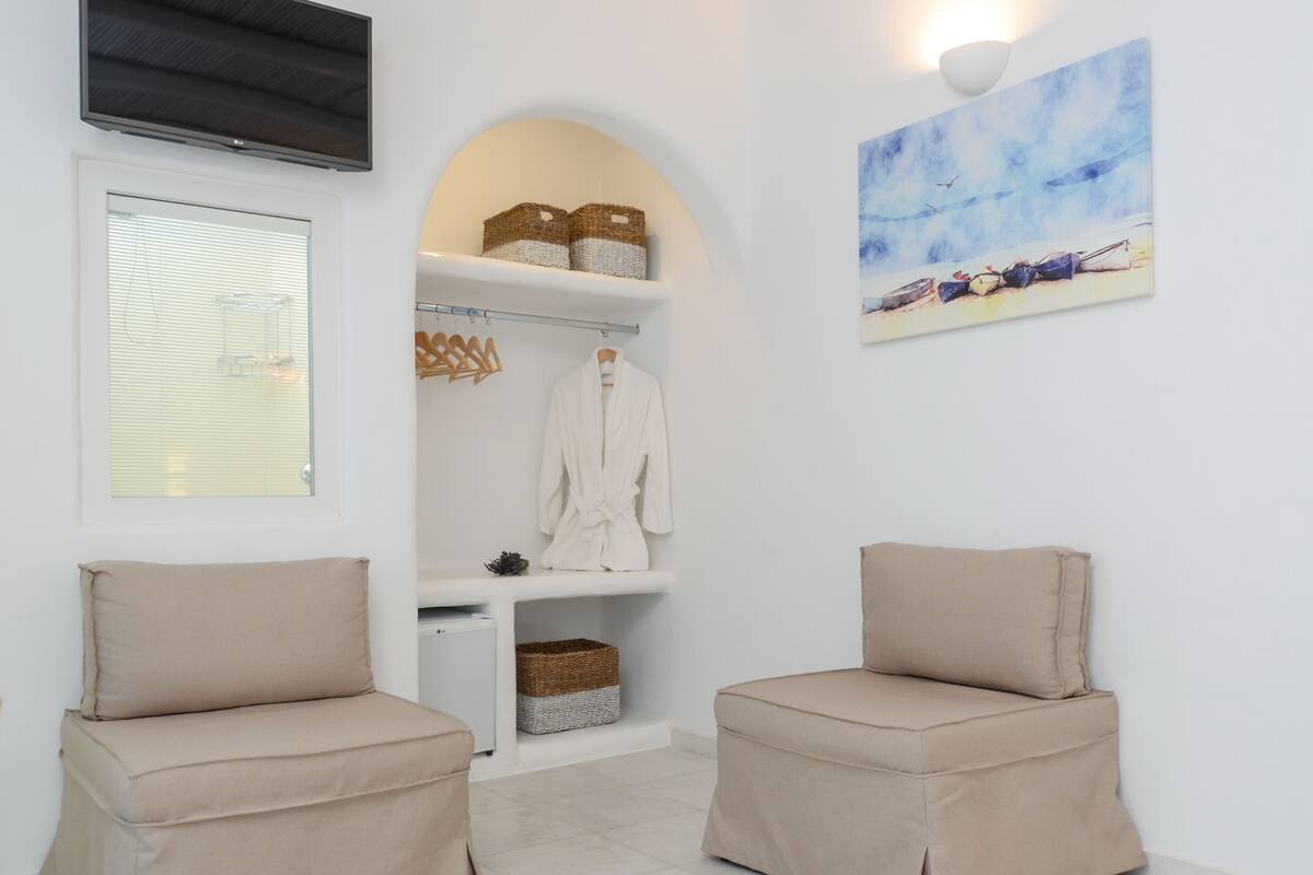 A spa room with two beige chairs, a white arched shelving unit with baskets and a robe, a painting of boats, and a TV mounted on the wall.