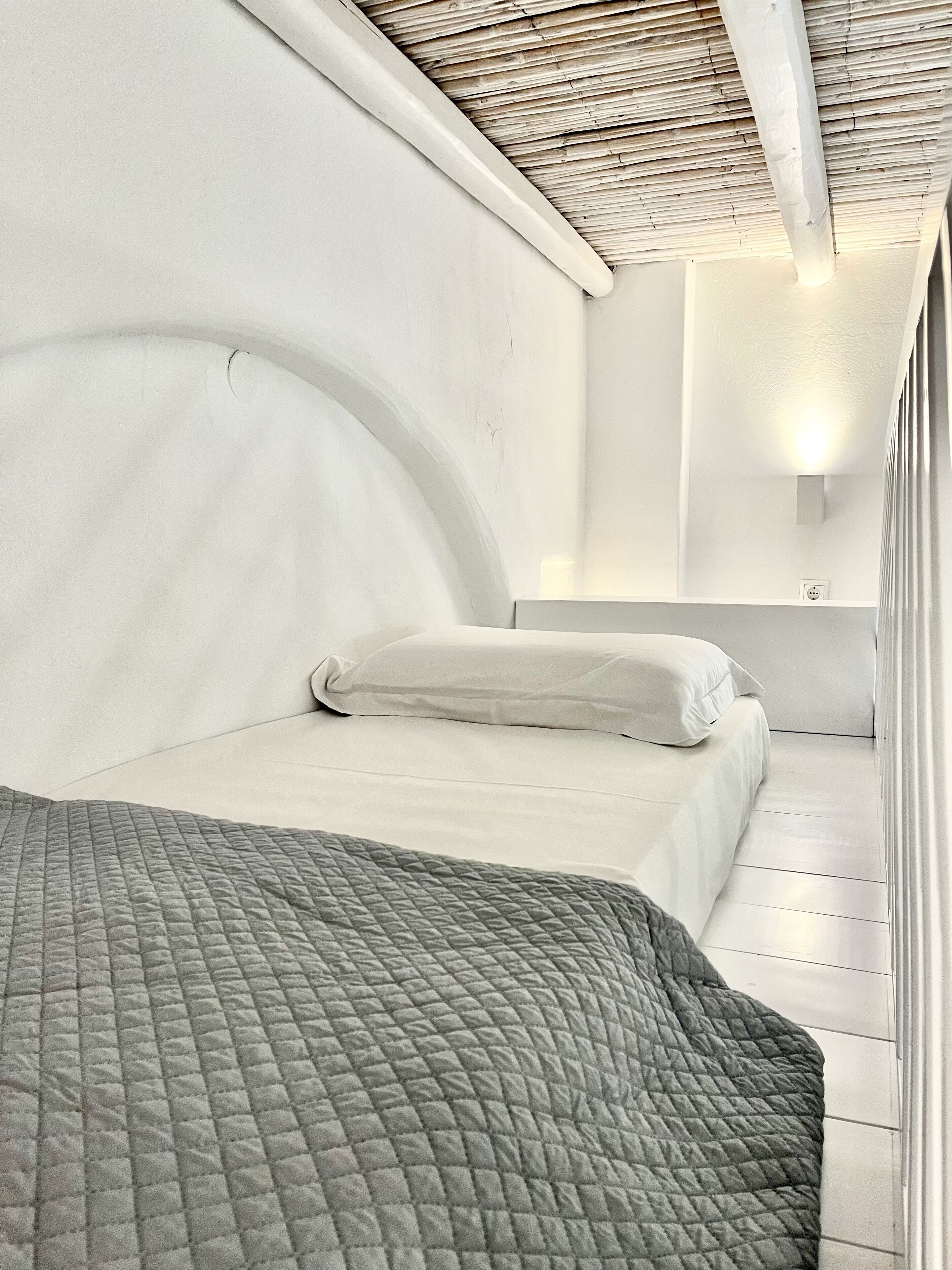 Ein modernes Schlafzimmer mit einem gewölbten Kopfteil, einem Bett mit einer gesteppten grauen Decke und einer weißen Wand mit einer Leuchte.