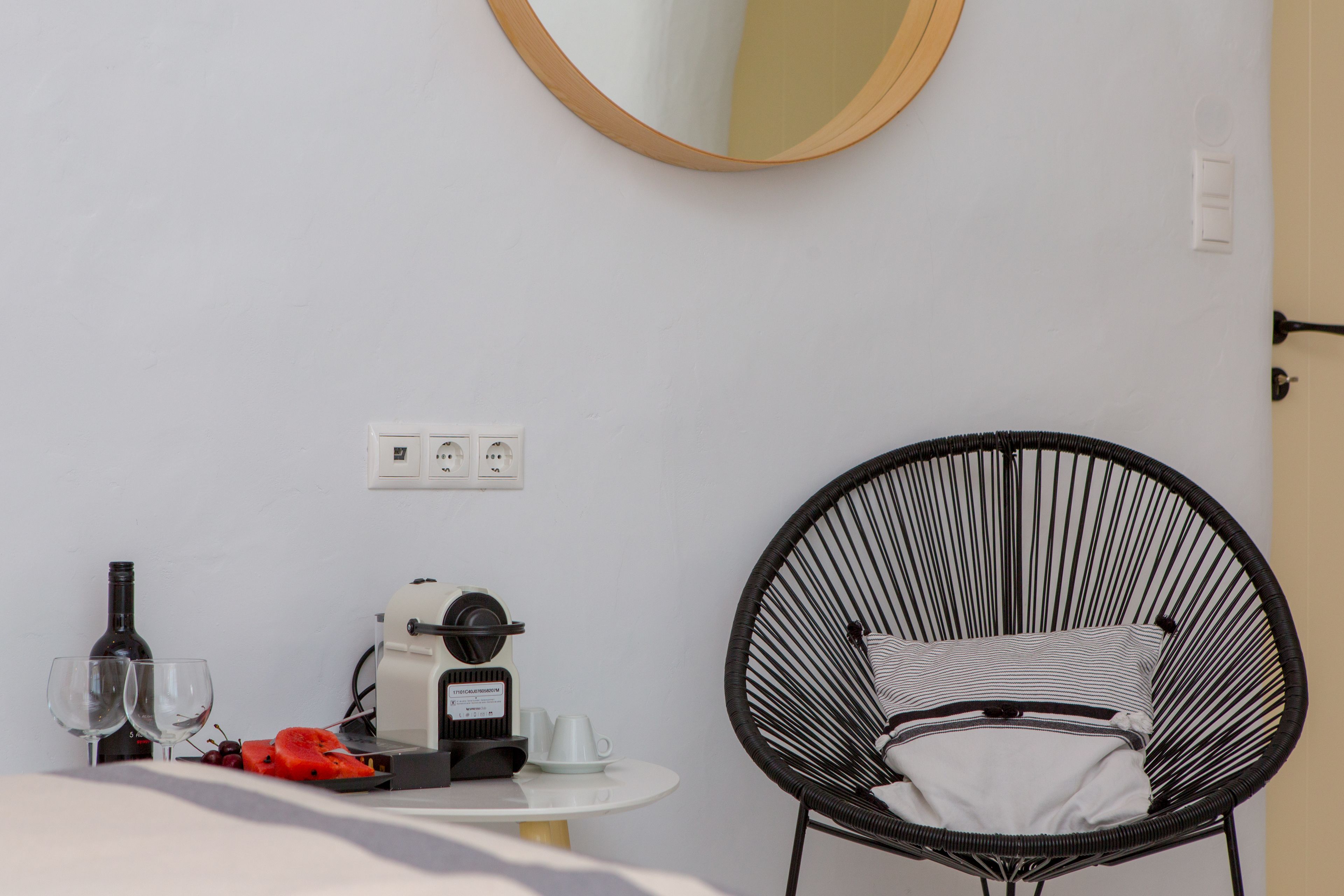 A hotel room with a round black chair, a small table with a coffee maker, wine glasses, and a bottle.