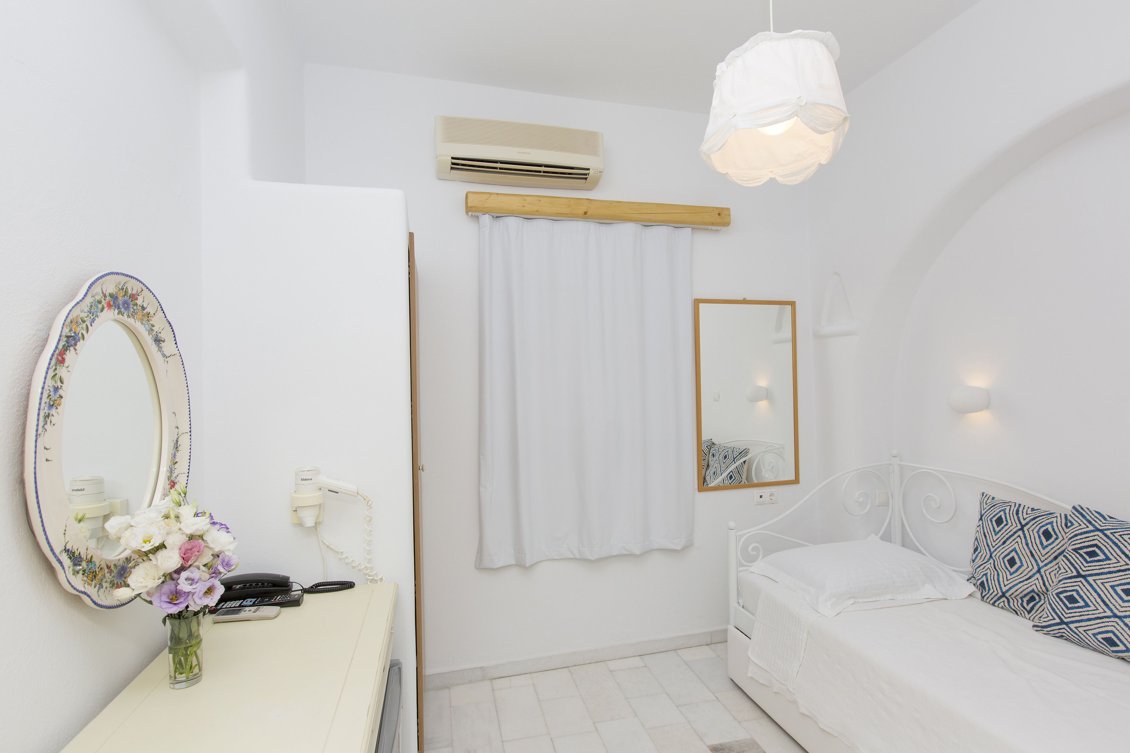 A white bedroom with a bed, a bedside table with a vase of flowers, a mirror, and a hanging light fixture.