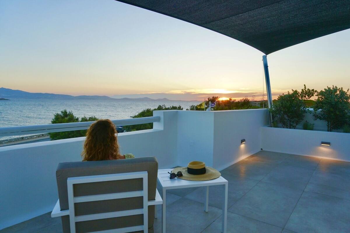 A person seated on a white chair on a balcony overlooking a sunset over the sea.