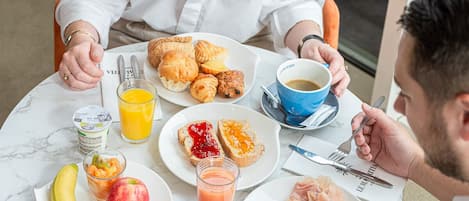 Una mesa redonda con alimentos para el desayuno, incluyendo cruasanes, tostadas, fruta y una taza de café.