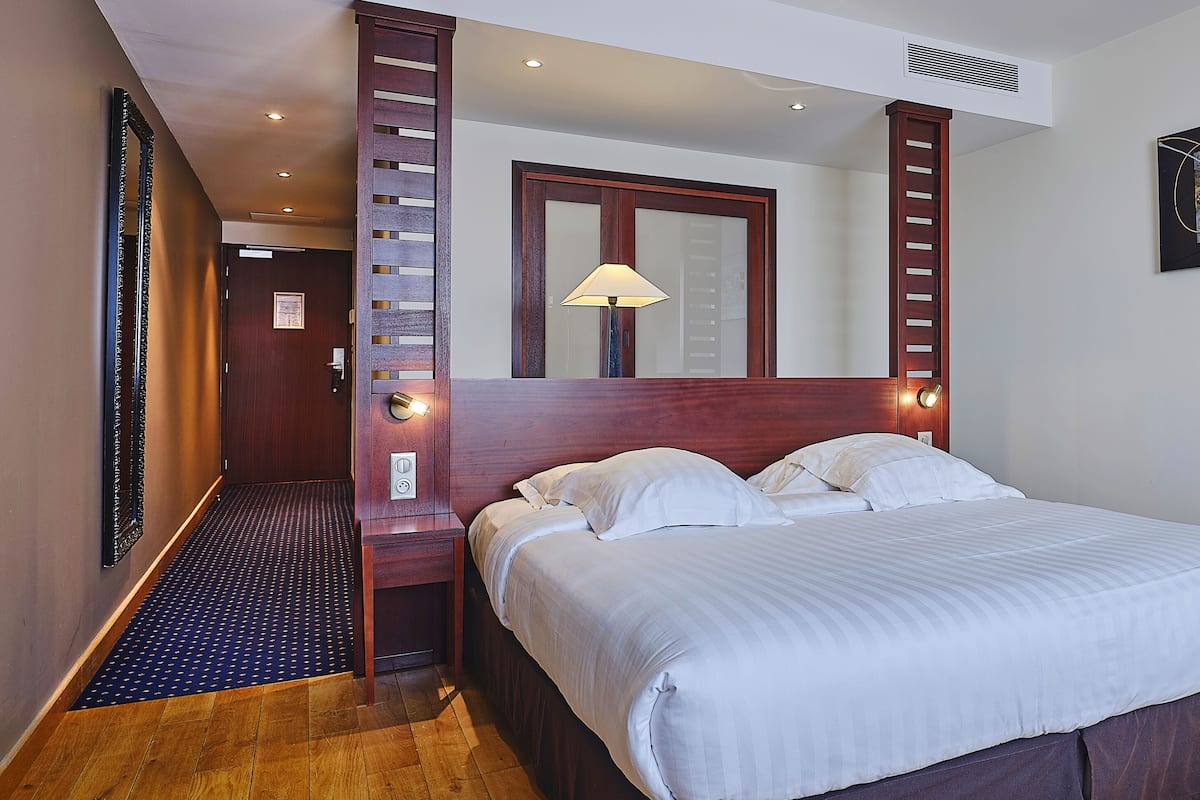 A hotel room with a large bed, wooden headboard, a nightstand with a lamp, a mirror, and a painting on the wall.
