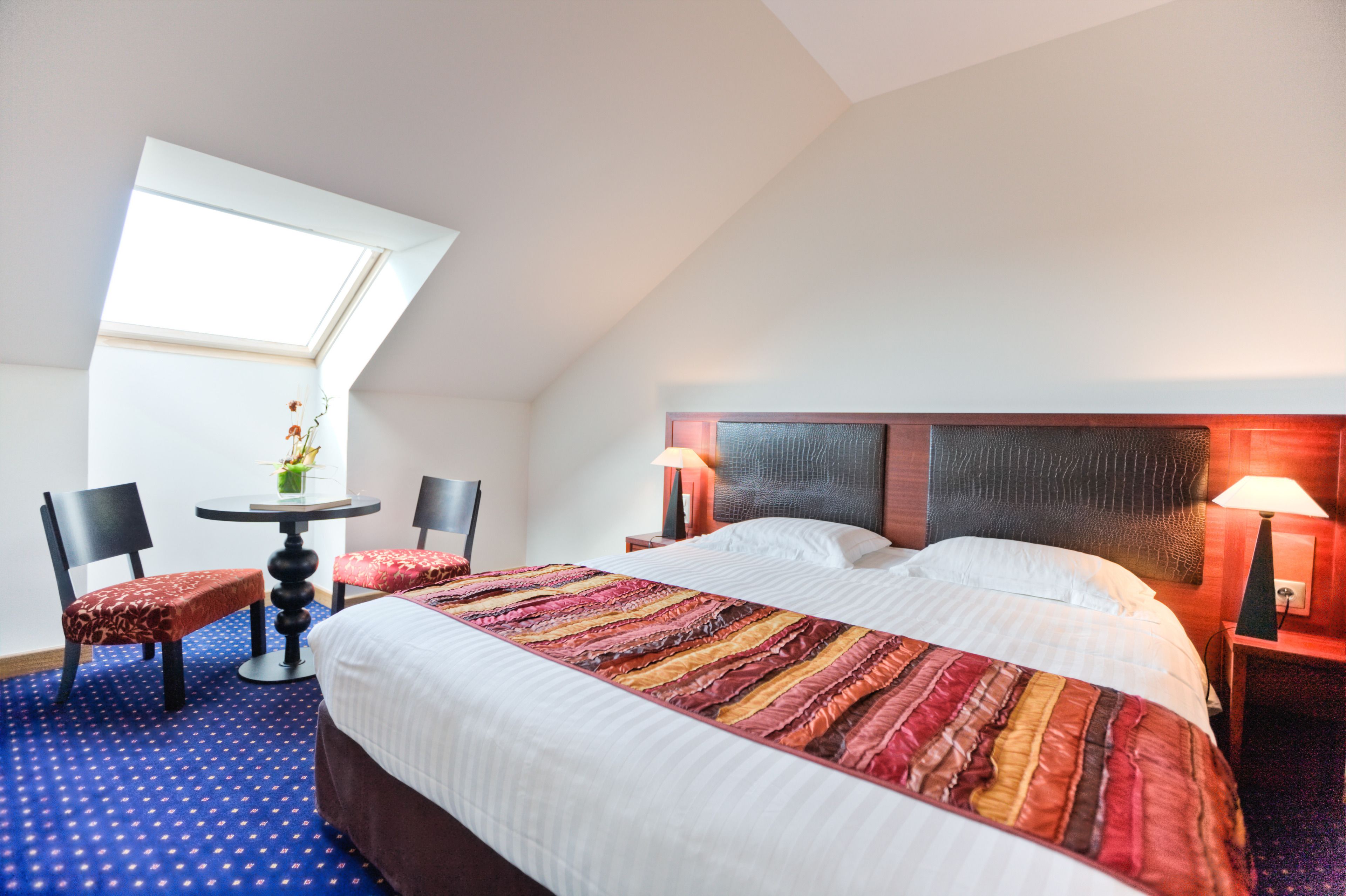 Una habitación de hotel con una cama grande, una mesita, dos sillas, un tragaluz y una mesita de noche con una lámpara.