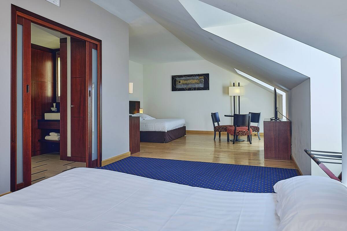 A hotel room with a large bed, a desk with a chair, a small table with two chairs, a lamp, and a painting on the wall.