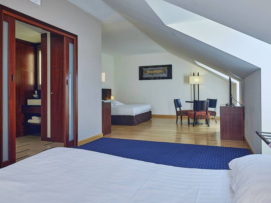 A hotel room with a large bed, a desk with a chair, a small table with two chairs, a lamp, and a painting on the wall.