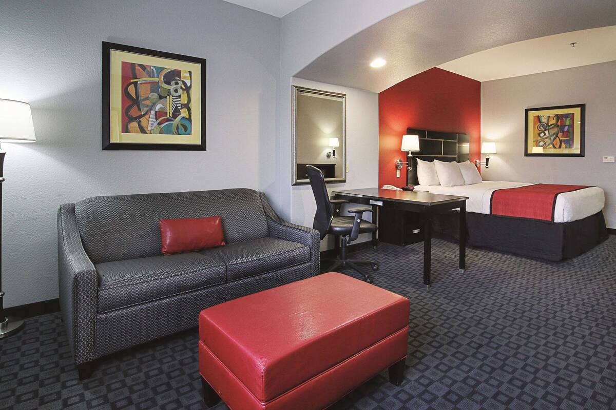 Habitación de hotel con sofá gris estampado, un otoman rojo, un escritorio pequeño, una cama con detalles en rojo y dos cuadros abstractos enmarcados en las paredes.