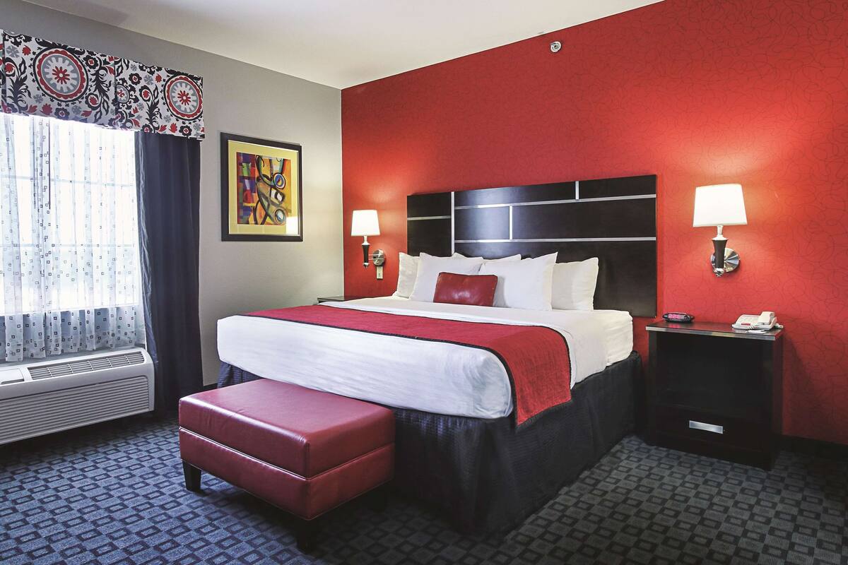 Habitación de hotel con una cama grande, un banco rojo, dos mesitas de noche con lámparas, una obra de arte enmarcada en la pared y una ventana con cortinas.