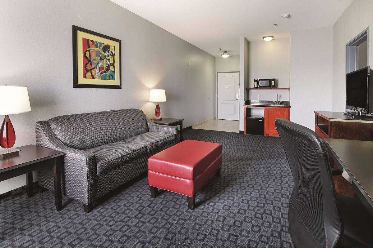 Habitación de hotel con alfombra gris estampada, sofá gris, reposapiés rojo, silla negra, mesita con lámpara, kitchenette con microondas y televisor, además de un cuadro abstracto enmarcado en la pared.